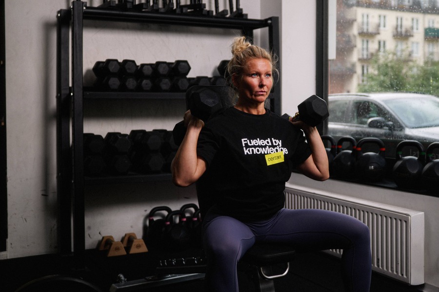 Kvinna som sitter på träningsbänk och kör axelpressar med hantlar